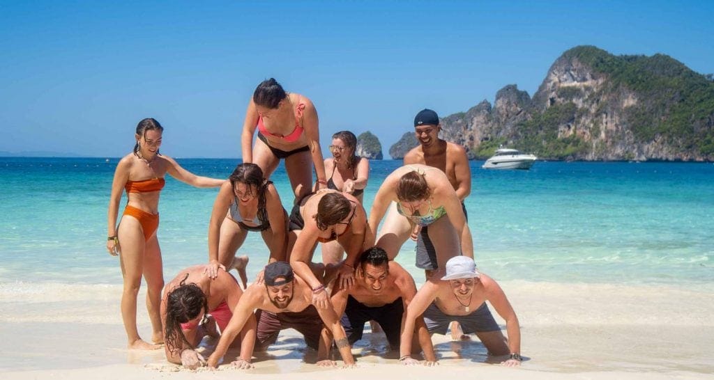 A joyful group of people engaged in building a human pyramid on a sandy beach with clear blue water and tropical mountains in the distance.
