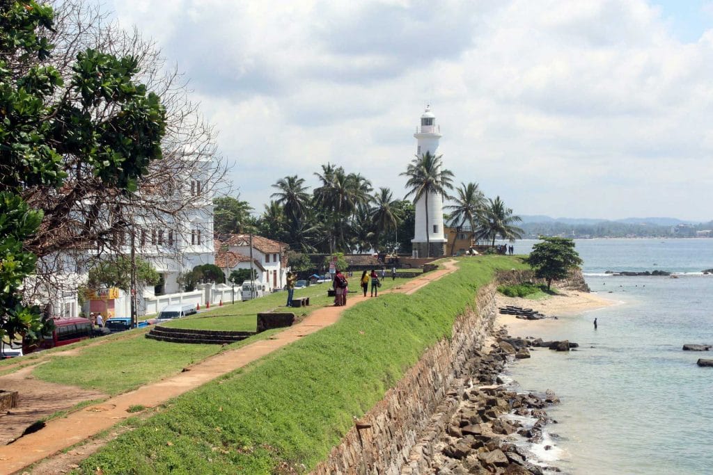 Scenic view of a coastal path with a white lighthouse to the right and ocean waves on the left, under a clear sky.

