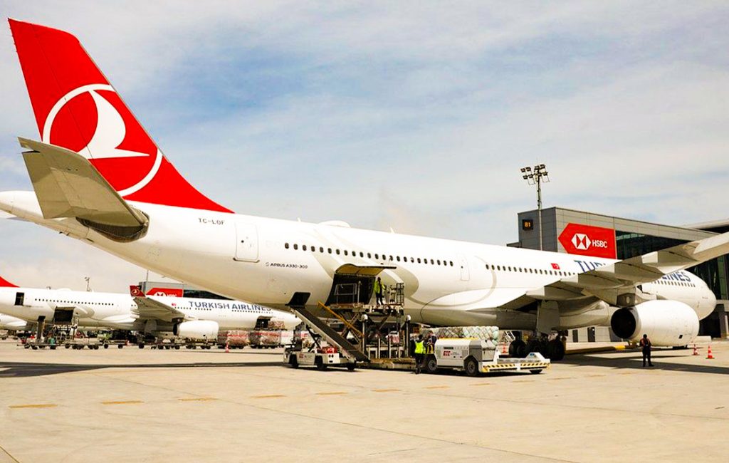 A Turkish Airlines Airbus A330-300 aircraft parked at an airport, being serviced. The plane is prominently displayed with other planes and airport infrastructure in the background