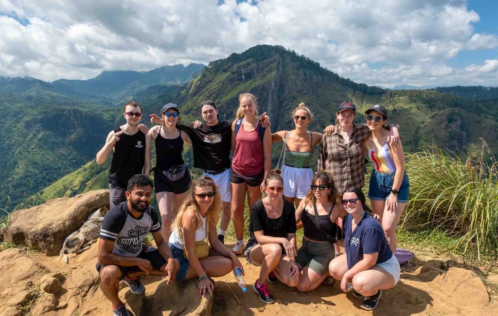 A group of eleven cheerful hikers posing for a photo with a panoramic view of a mountainous landscape, under a clear blue sky