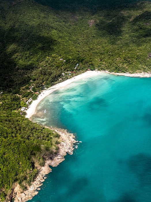 Aerial view of a serene, crescent-shaped beach in Thailand with turquoise waters, bordered by a lush green forest and rocky headlands.