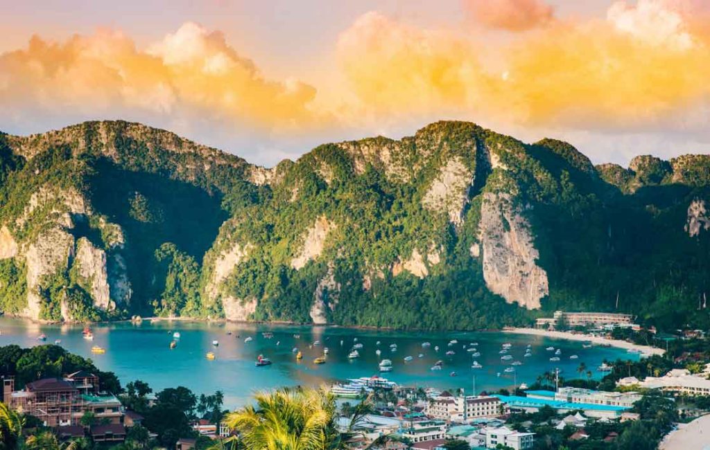 Overlook of a tropical bay with boats anchored in the water and a backdrop of mountainous terrain under a sunset sky