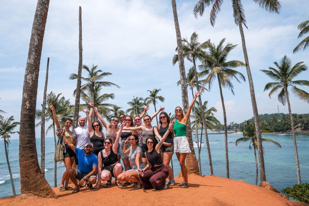 A group of diverse friends posing with arms raised, celebrating amidst tall coconut trees with a scenic view of the ocean and clear skies in the background. Out of the pros and cons, one of the biggest pros is the instant group of friends.