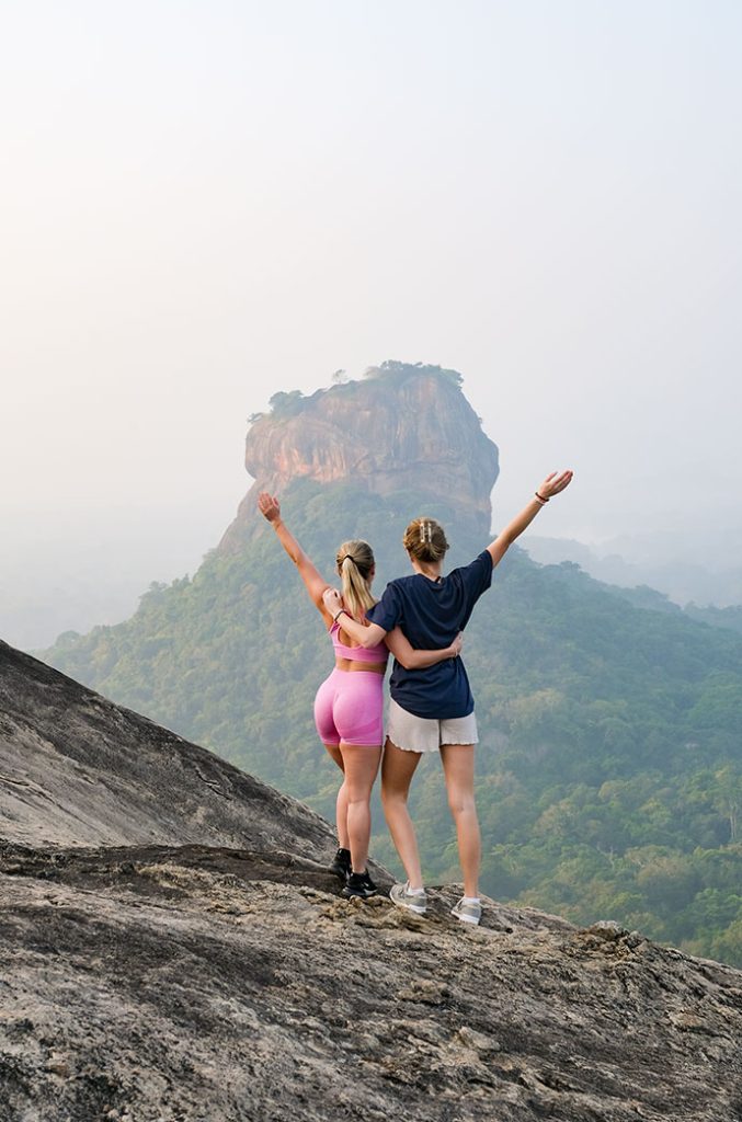 Two travelers with arms raised in triumph, overlooking a stunning mountainous landscape from a high vantage point.