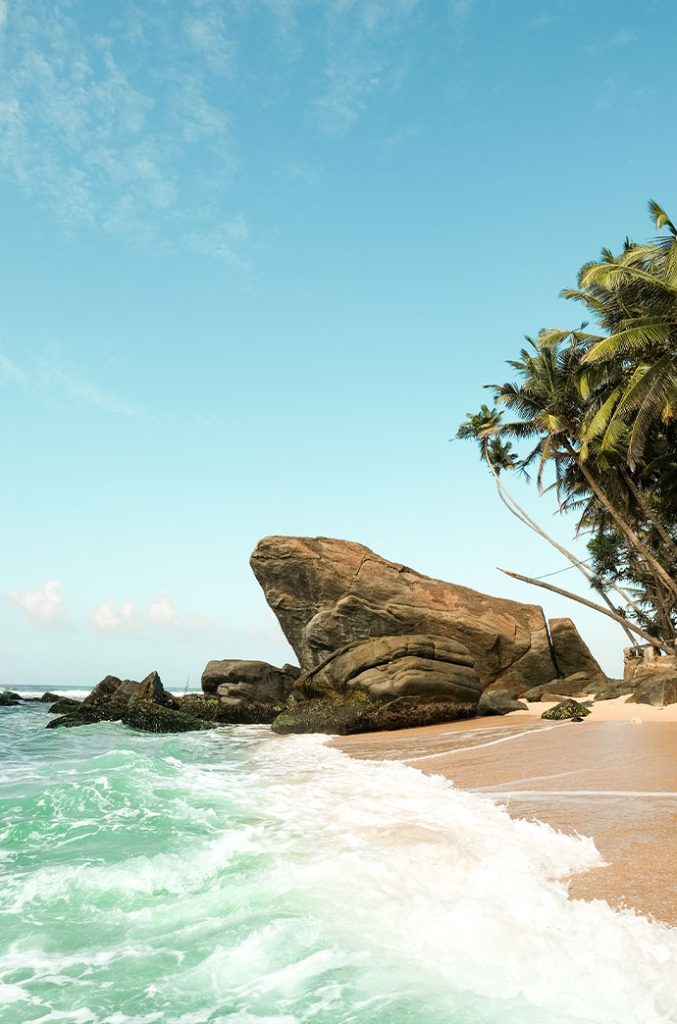 A vivid beachscape with a large, smooth rock formation on sandy shores washed by foamy waves, under a clear sky with scattered clouds