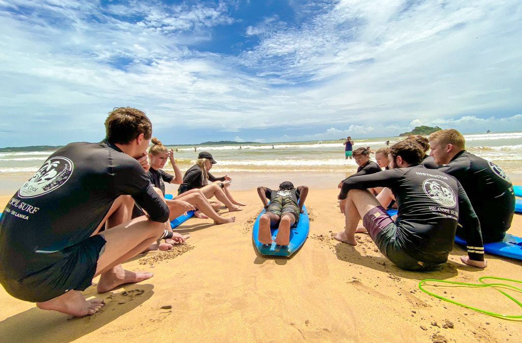 A surfing lesson in session on a beach, with a group of people seated on surfboards listening to an instructor, ocean waves in the background.
