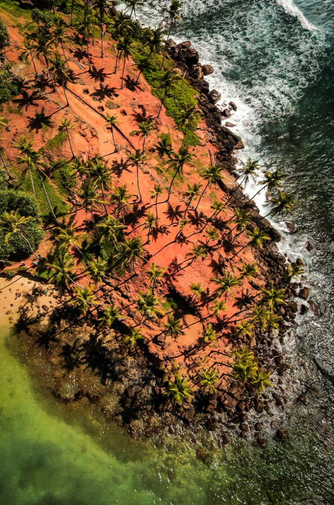 Bird's-eye view of a red-earth peninsula with palm trees, bordered by rough sea on one side and calm waters on the other