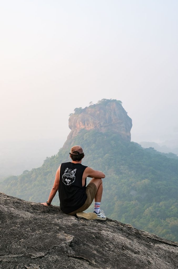 A solitary figure sits contemplatively on a mountain edge, gazing out at a distant rock fortress amidst a sea of greenery.