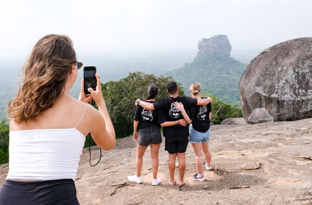 A woman taking a photo with her smartphone of three people looking at a large rock formation and green forest from a mountaintop. One of the biggest pros and cons of group travel is taking photos.
