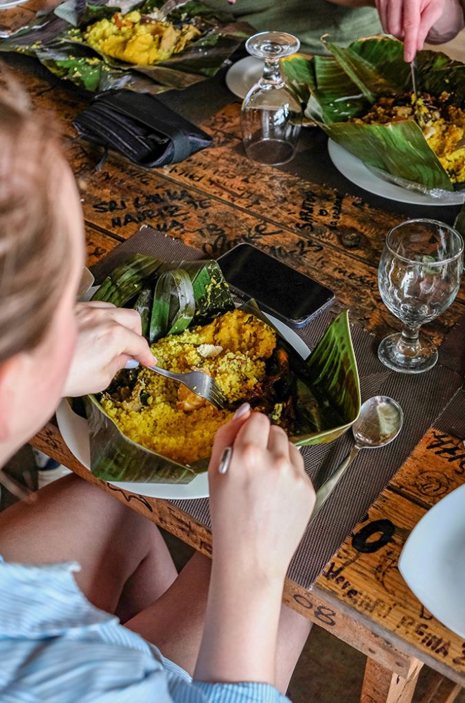 A person seated at a wooden table, eating a traditional meal wrapped in banana leaves at a rustic dining setting