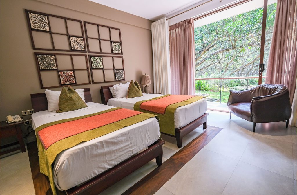 A modern, well-lit twin bedroom with a large window overlooking a forest, decorated with colorful bedding and a collection of square patterned frames on the wall.