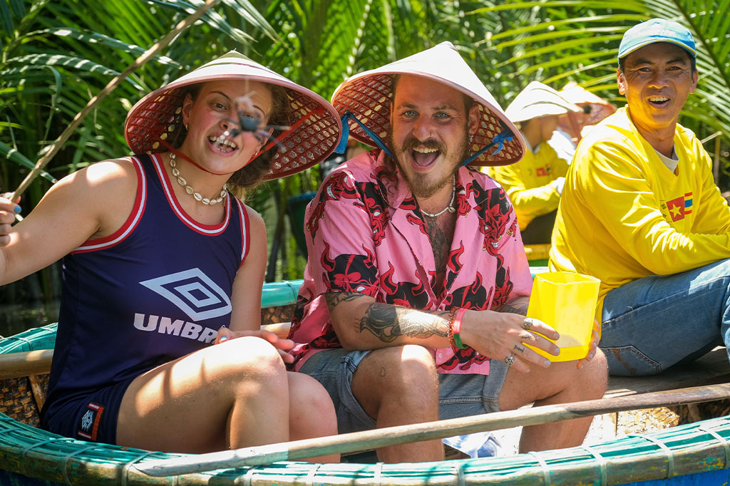 Two tourists, wearing traditional Vietnamese conical hats, smile brightly while sitting in a basket boat, likely on a fishing tour in Hanoi. One holds a fishing rod, while the other holds a yellow plastic cup, surrounded by lush green palm trees in the background. A local guide, also wearing a hat and bright yellow shirt, sits behind them, adding to the lively atmosphere of the outdoor adventure.