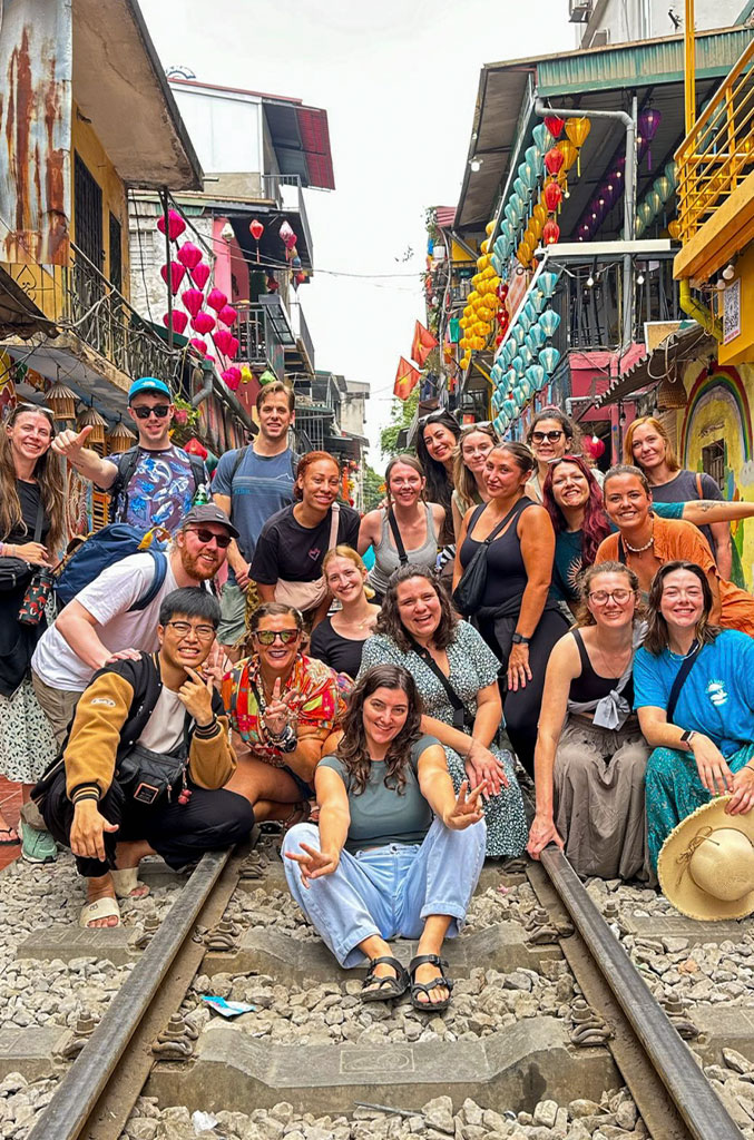 A large group of tourists poses happily on the famous Hanoi Train Street, sitting and standing between the railway tracks. Behind them, colorful lanterns and vibrant buildings line the narrow street, adding a festive atmosphere. The group smiles and flashes peace signs, capturing a fun and lively moment on this unique urban street.