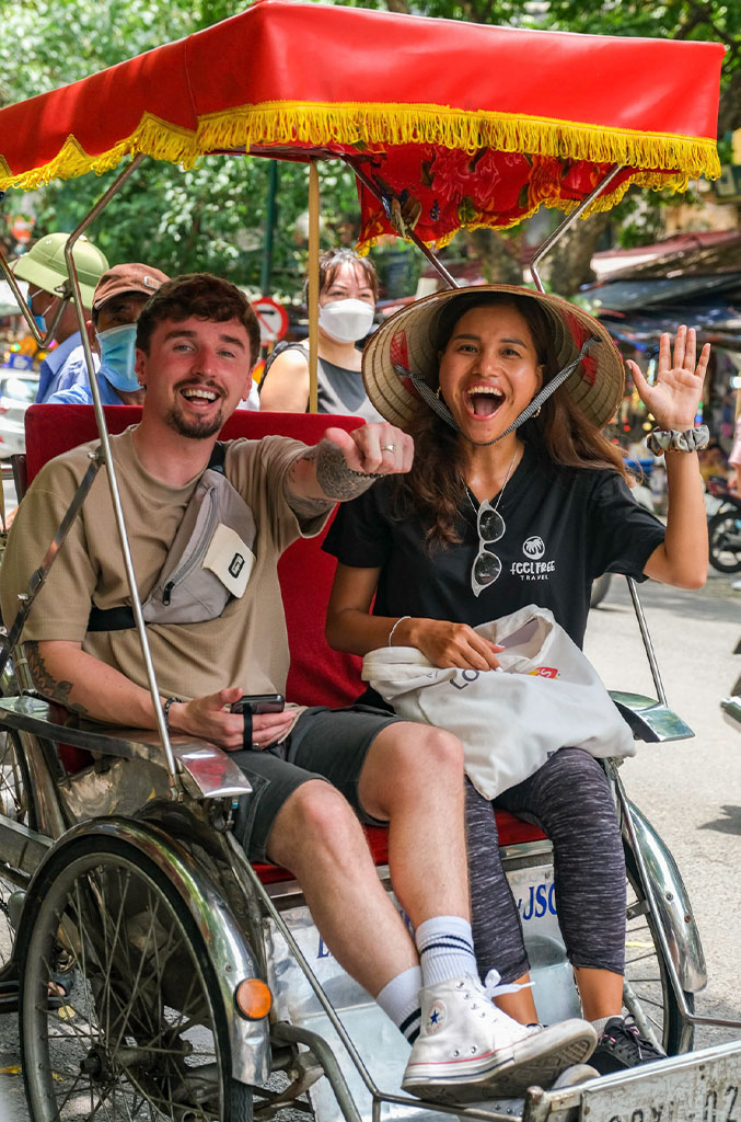 Two smiling people sit in a vibrant red and yellow rickshaw, likely in Hanoi. One person is pointing excitedly while the other waves with joy, wearing a traditional Vietnamese conical hat. Both appear to be enjoying a lively street scene, surrounded by pedestrians and greenery.