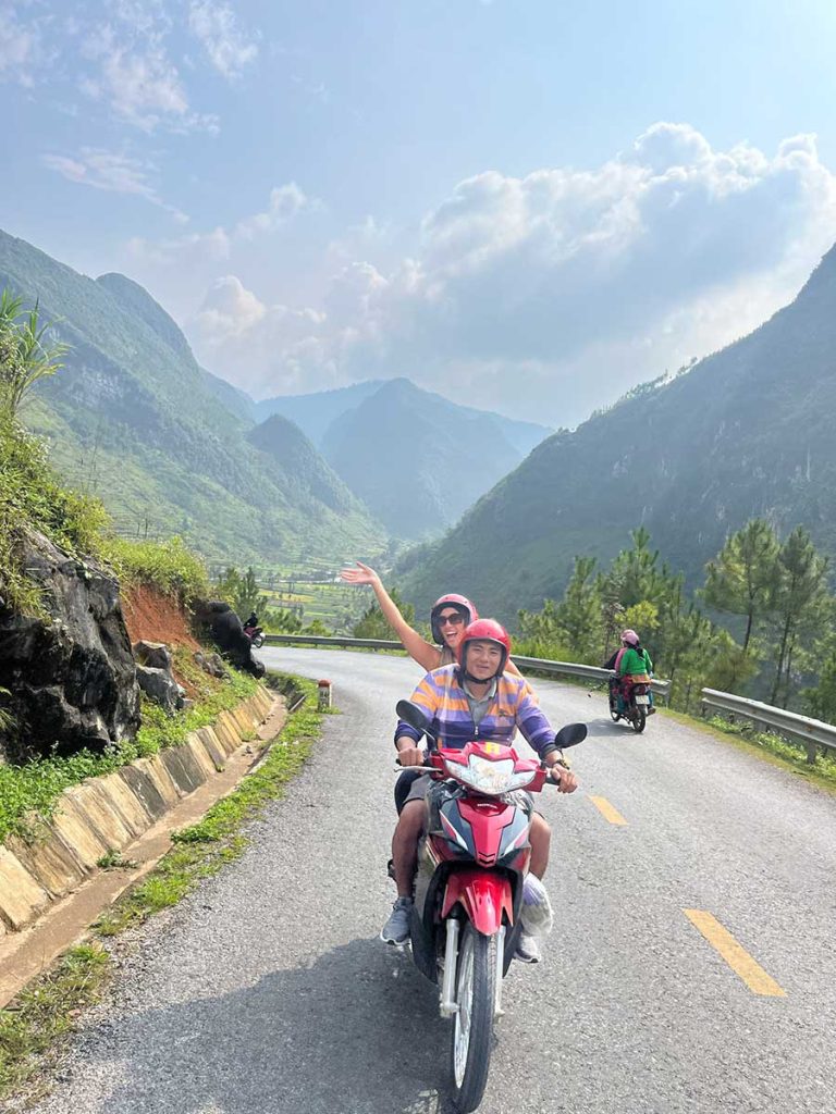 A picture of a traveller on the back of a bike with an EasyRider. She is smiling and waving at the camera. 