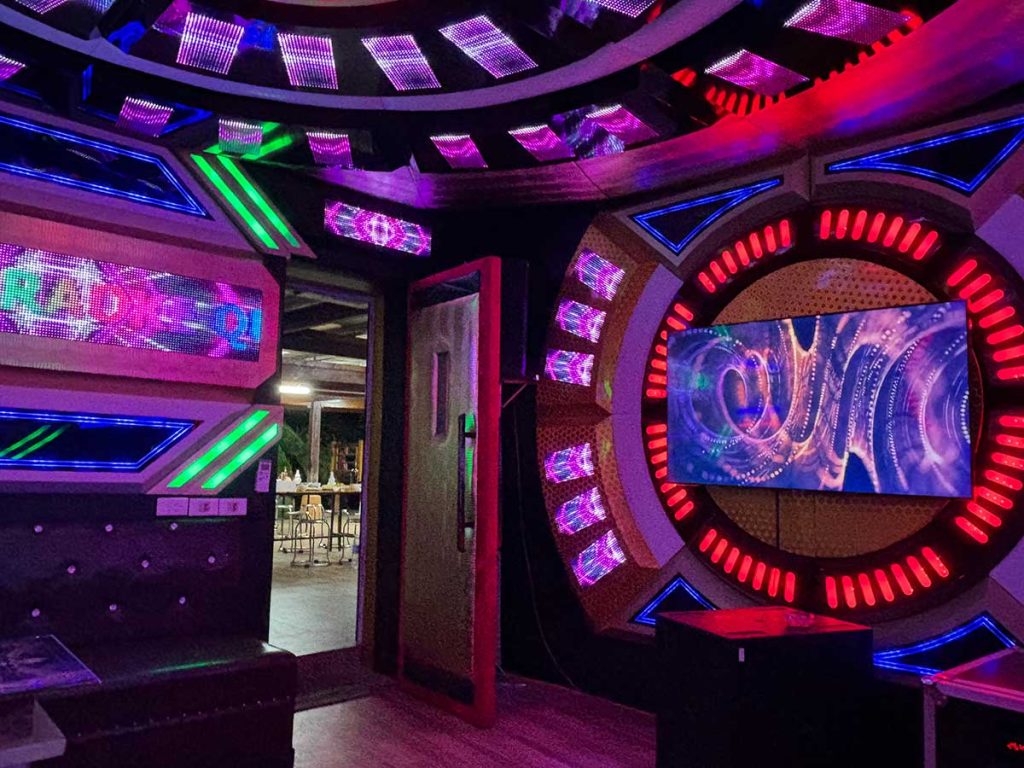 A small, cozy karaoke room with vibrant, colorful lighting. Microphones and a screen are set up in front of cushioned seating, inviting guests to sing and enjoy the lively atmosphere.