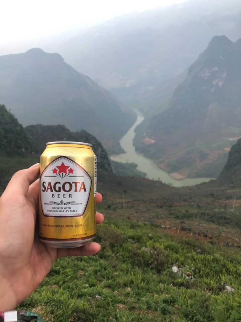 A glass of beer placed on a wooden table, with mountains and valleys visible in the background. The golden beer is frothy, and the setting suggests a relaxed moment after a day of exploration.