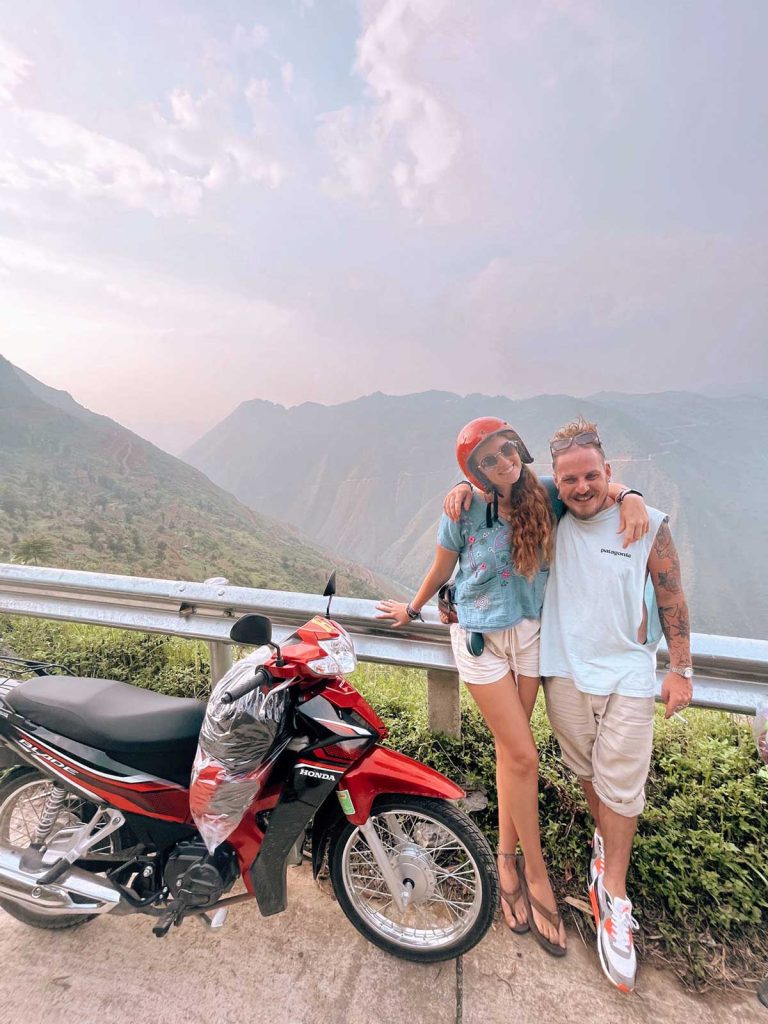 A couple posing by their bike along the Ha Giang Loop with a stunning mountain range in the background