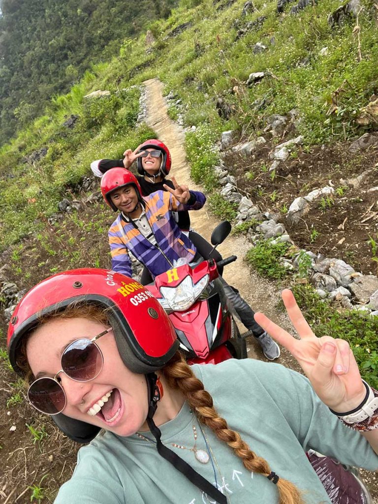 A group of travelers taking a selfie on a mountain road, surrounded by breathtaking views of valleys and peaks. They are smiling and seem to be enjoying the adventure, with the expansive landscape behind them.