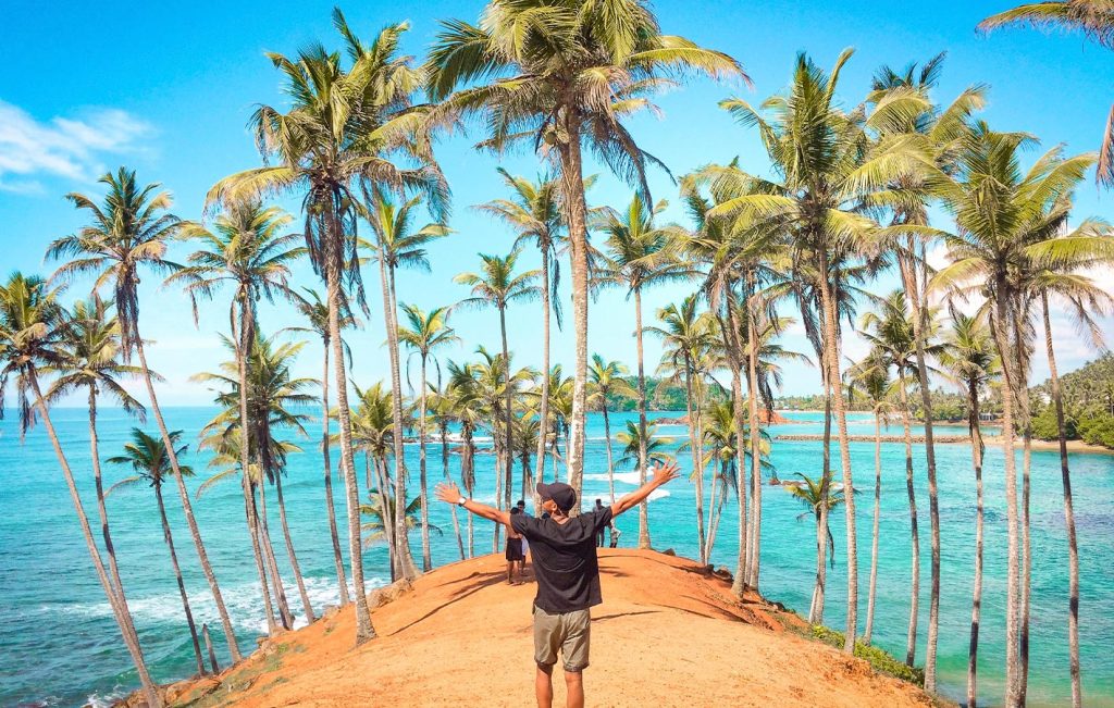 A traveler standing on Coconut Tree Hill in Mirissa, Sri Lanka, overlooking turquoise waters and palm trees.