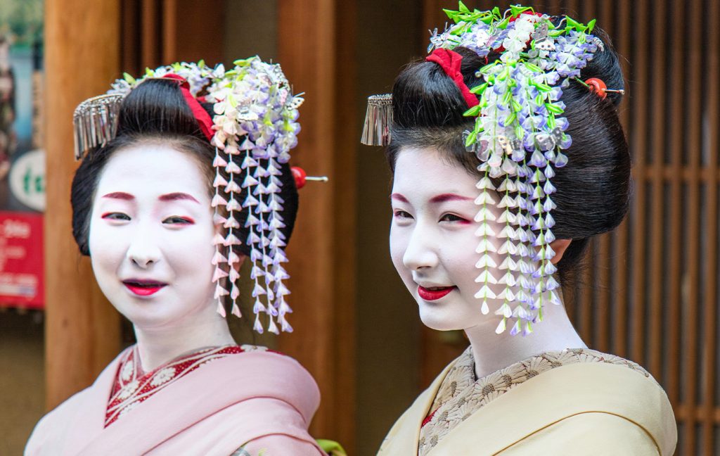 Two women dressed as geisha with white makeup and colorful hair ornaments smile in traditional attire.