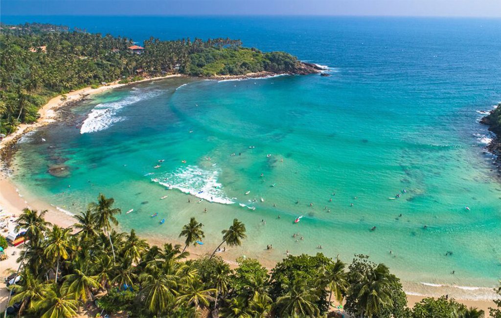 A panoramic aerial view of Weligama Bay with surfers enjoying the clear blue waves.

