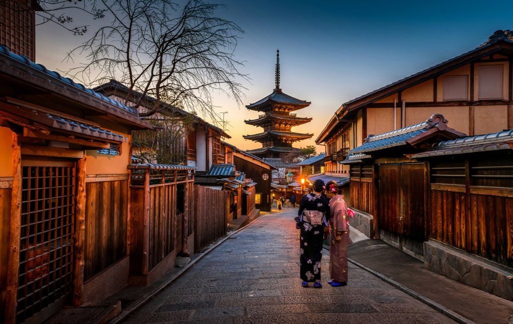 Two women in kimonos stand on a traditional street in Kyoto with Yasaka Pagoda in the background at sunset.