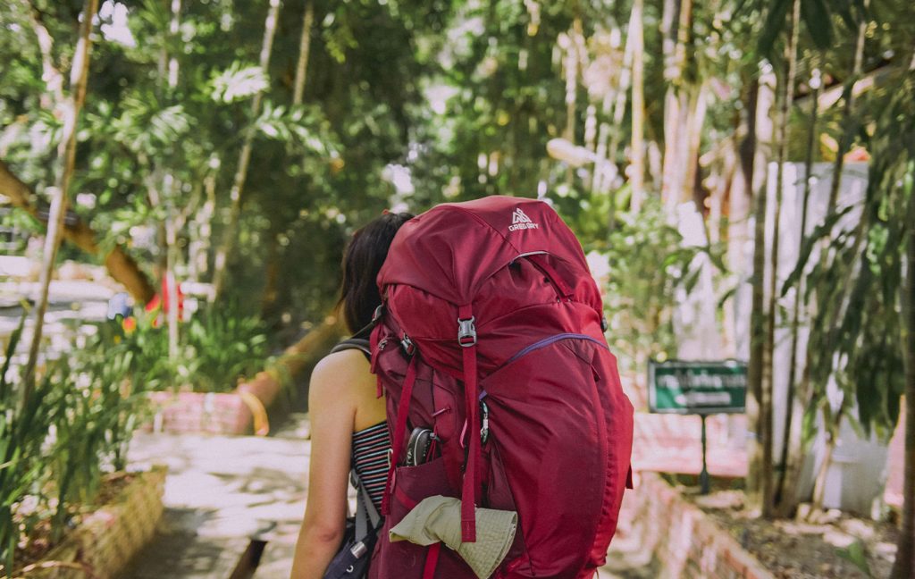 A traveler with a large red backpack walks along a sunny garden path.