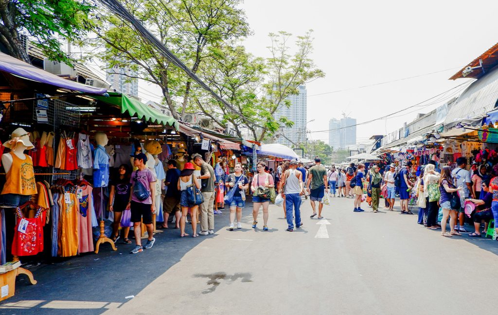 A busy outdoor market is lined with clothing stalls and crowds of shoppers.
