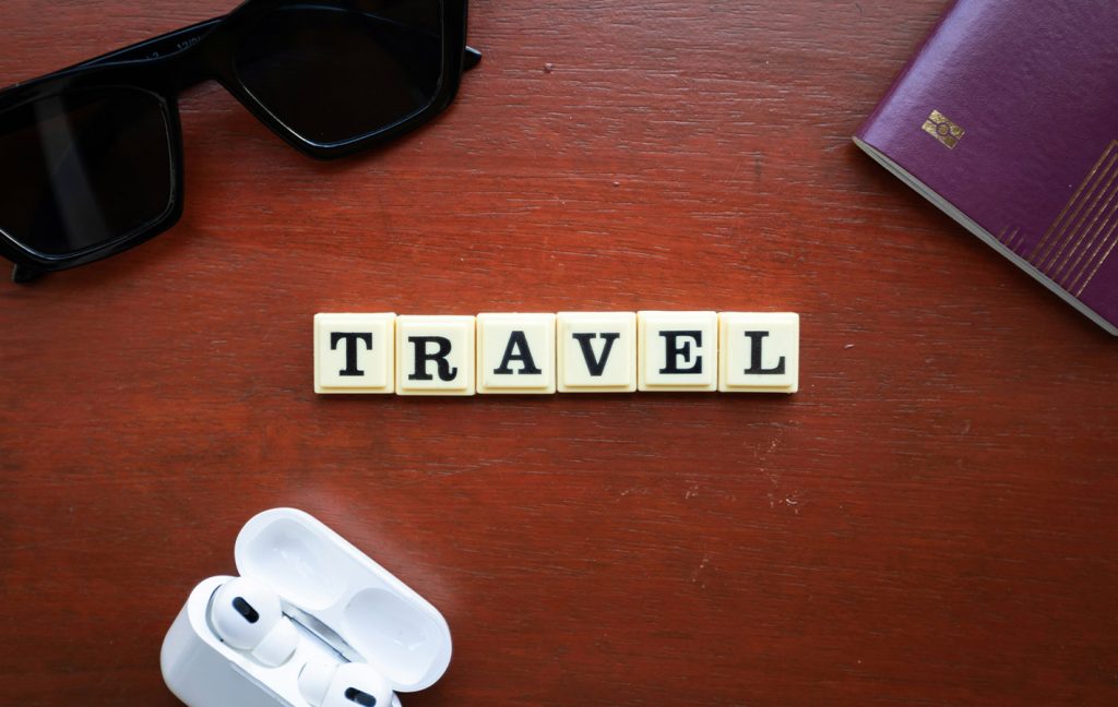 Wooden tiles spelling “TRAVEL” sit on a table beside sunglasses, earphones, and a passport.
