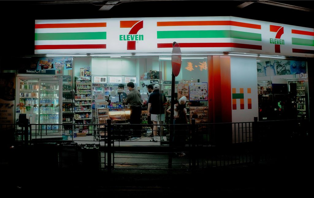 A brightly lit 7-Eleven store is seen at night with customers inside.