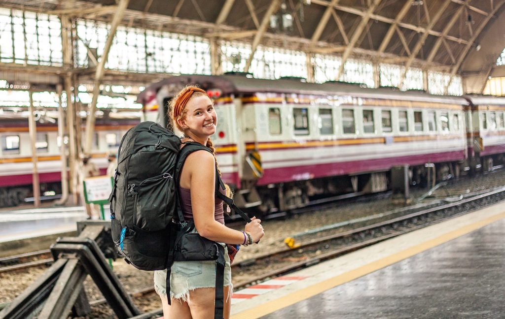 A smiling backpacker stands on a train platform beside a stationary train.