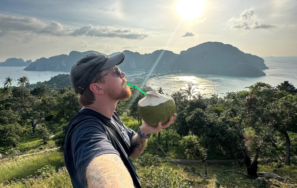 A man drinks from a fresh coconut while overlooking a tropical island landscape.