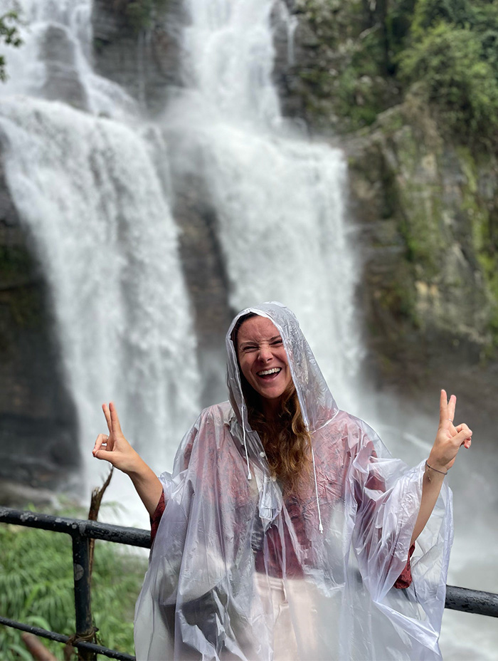 A smiling woman in a poncho flashes peace signs in front of a powerful waterfall.