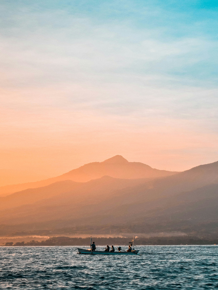 A small boat with people on board sails across the ocean at sunrise with mountains in the background.