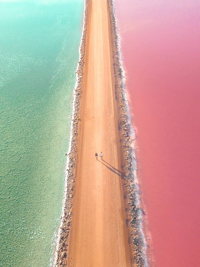 A striking pink and green lake divided by a narrow dirt road with two people walking.