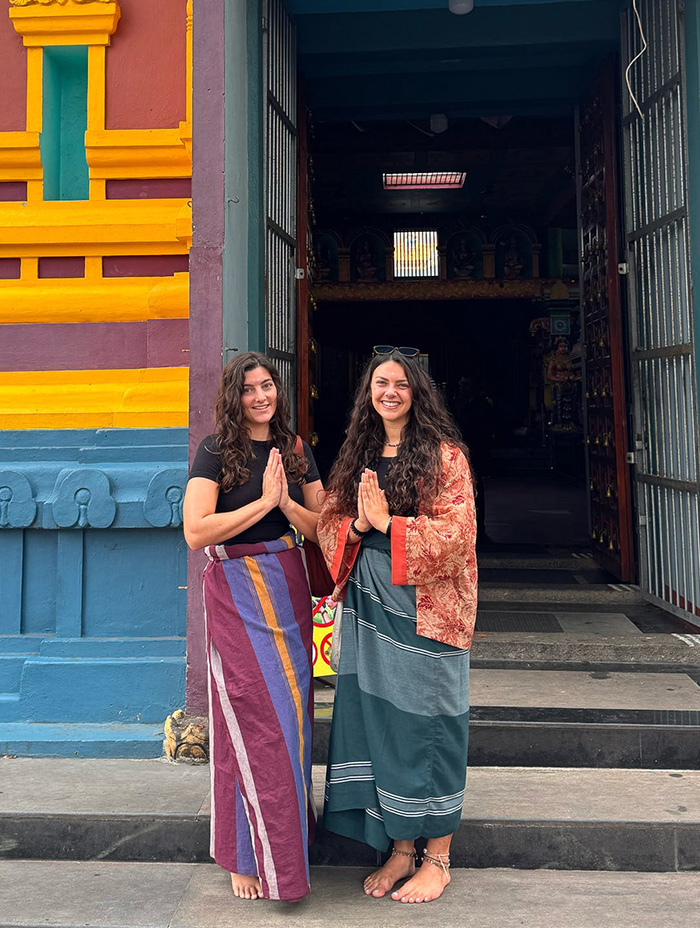 Two women wearing sarongs pose outside a colorful temple entrance.