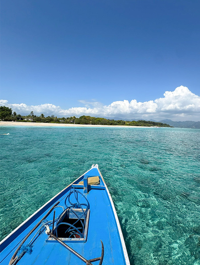 The bow of a blue boat points toward a tropical shoreline over clear turquoise water.