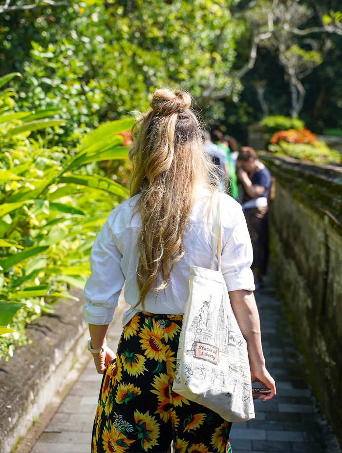 A woman with long blonde hair and a tote bag walks down a lush garden path.