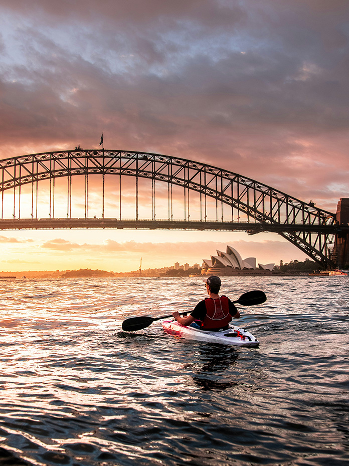 A kayaker paddles on Sydney Harbour with the Harbour Bridge and Opera House in view.