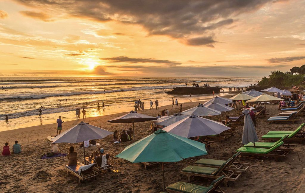 A crowded beach in Bali at sunset with umbrellas, loungers, and people enjoying the view.