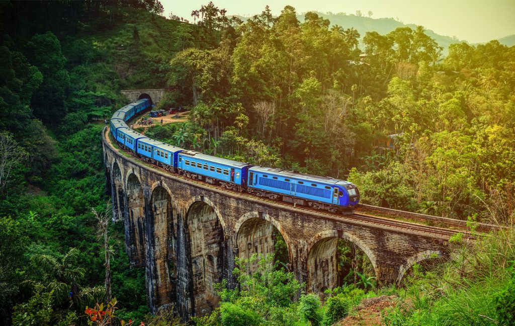 A blue train travels across the iconic Nine Arches Bridge in lush Sri Lankan hills.