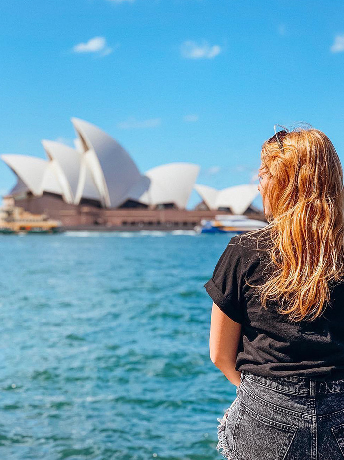 A woman with blonde hair admires the Sydney Opera House from across the harbor.