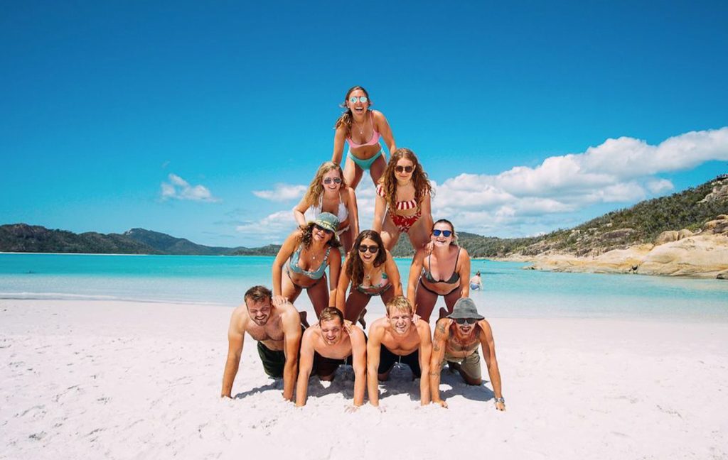 A group of friends form a human pyramid on a bright sandy beach.