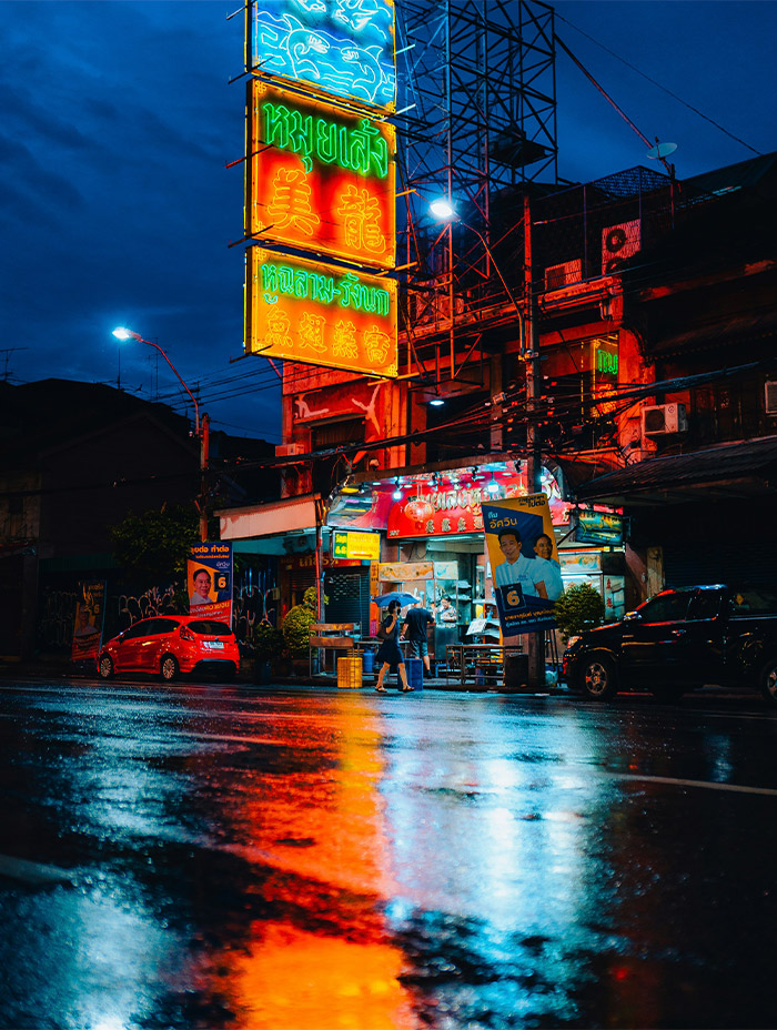 Neon signs reflect on wet pavement outside a street food shop at night.