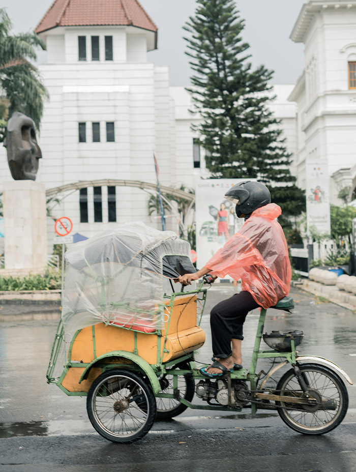 A man in a rain poncho rides a tricycle taxi along a wet street.