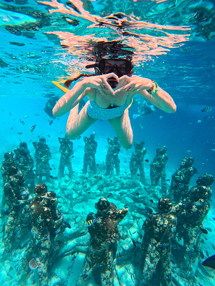 A snorkeler forms a heart shape with her hands above an underwater sculpture circle.