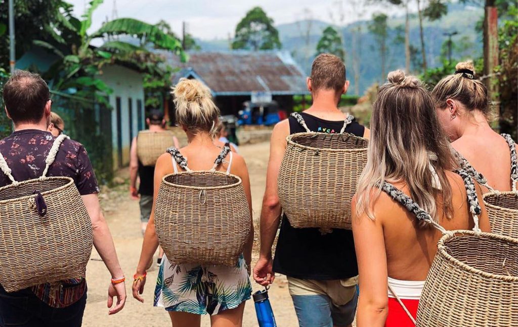 A group of travelers walk through a village carrying large woven baskets on their backs.