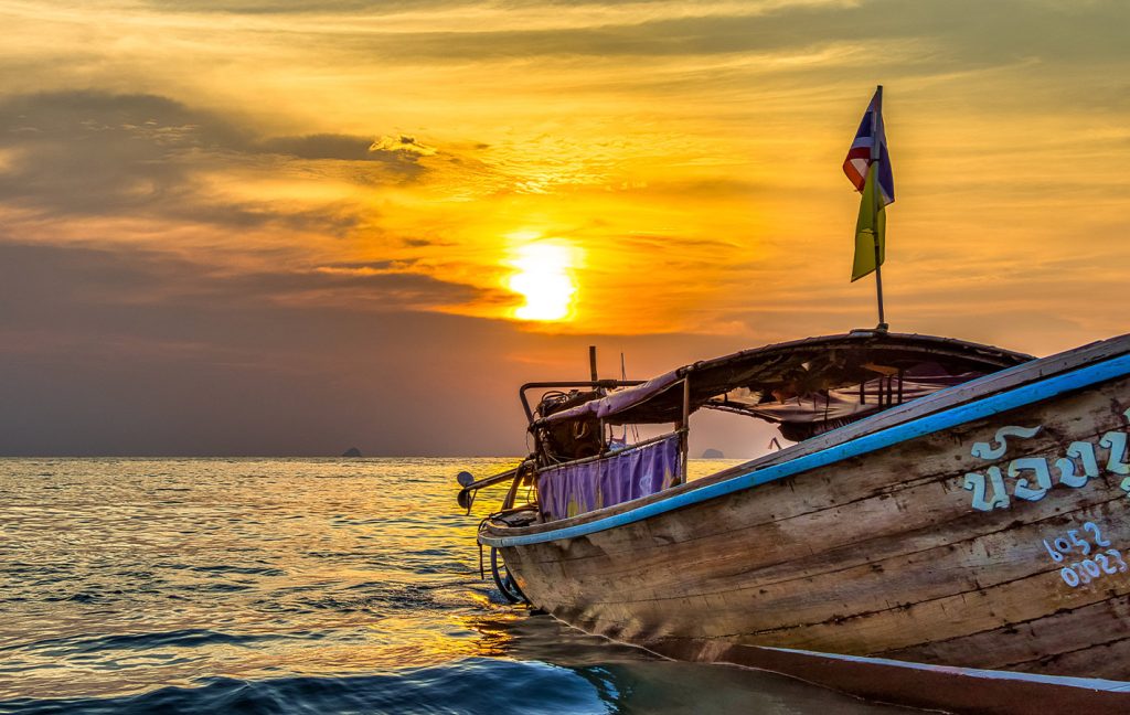 A wooden longtail boat floats on calm water during a golden sunset.