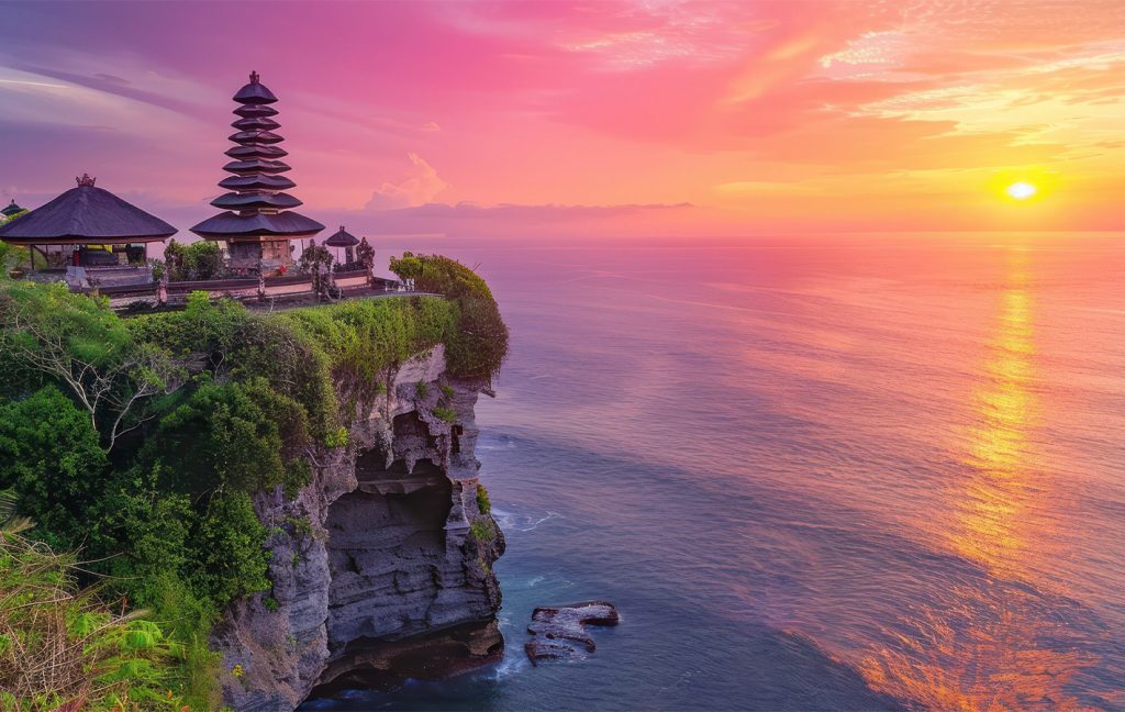 A cliffside temple in Bali overlooks the ocean at sunset with a pink and orange sky.